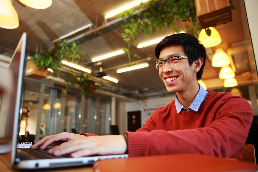 Cheerful Asian man wearing glasses typing on a laptop in a cafe