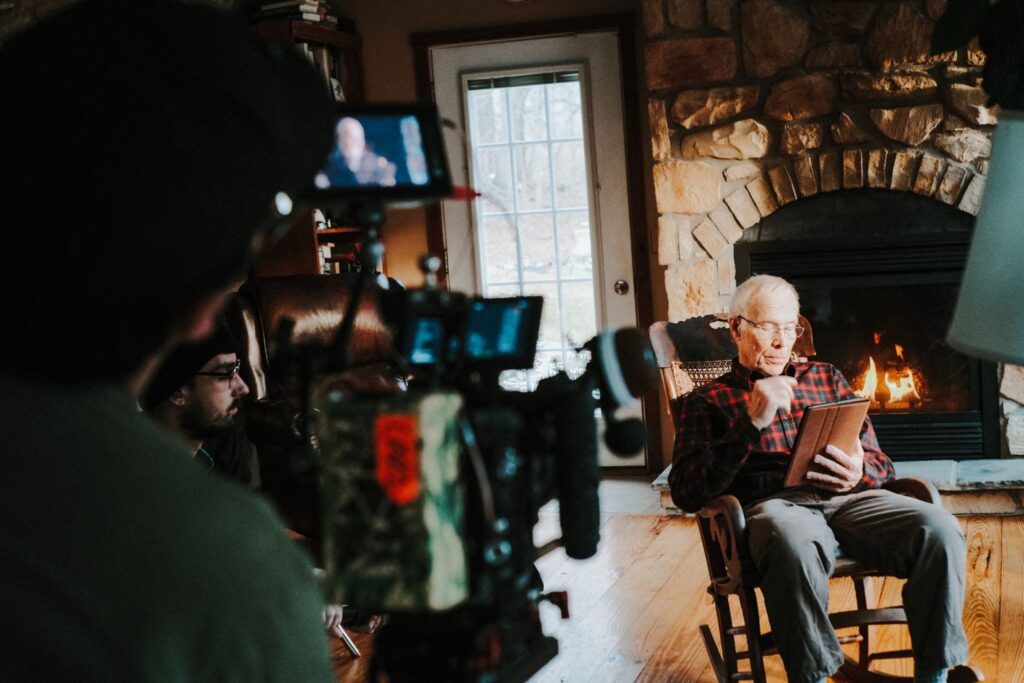 Over the shoulder image of an elderly man sitting in a rocking chair in front of a fireplace looking at a tablet while film professionals record him with large film cameras